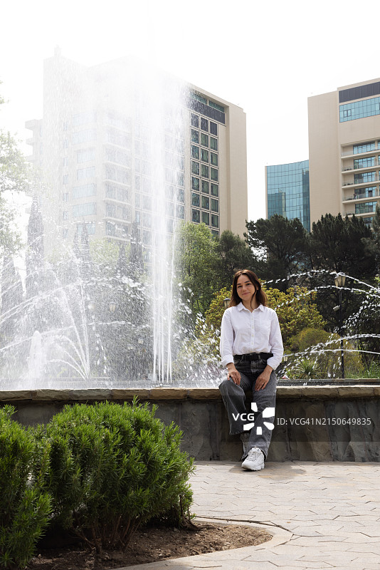 一位穿着牛仔裤和白衬衫的年轻漂亮黑发女孩走在街上，身后是高大的混凝土建筑和喷泉。图片素材