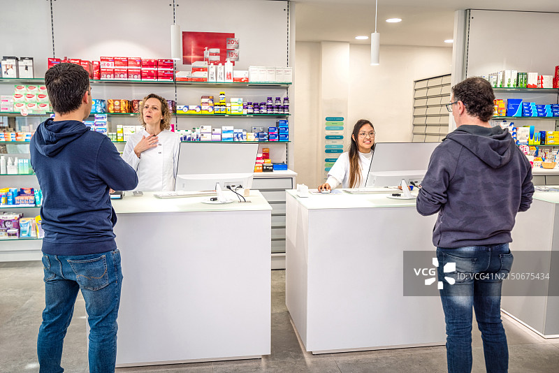 现代药房，货架上摆满了现代药品、药物、维生素盒、补品。背景是两位专业药剂师在收银台为两位顾客服务图片素材