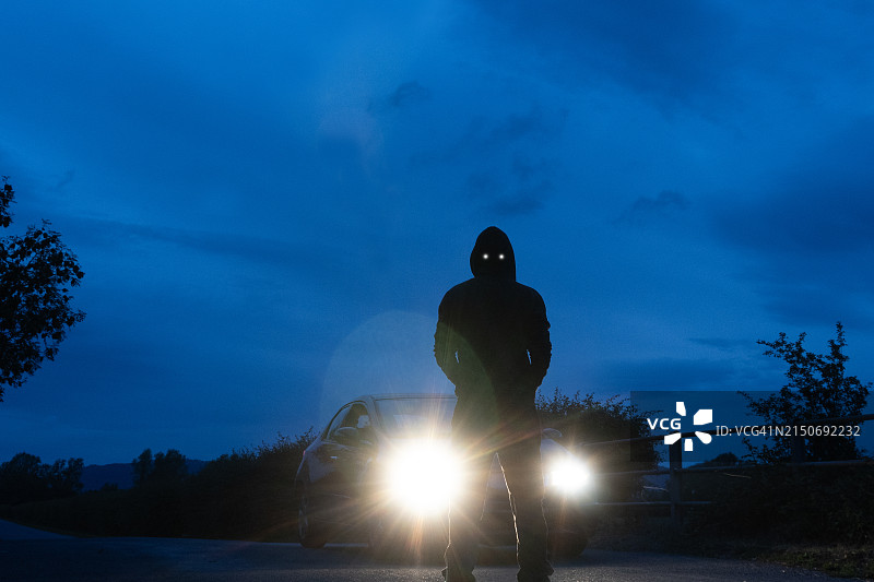 超自然恶魔在开启大灯的汽车前，夜晚乡村公路上的恐怖概念图片素材