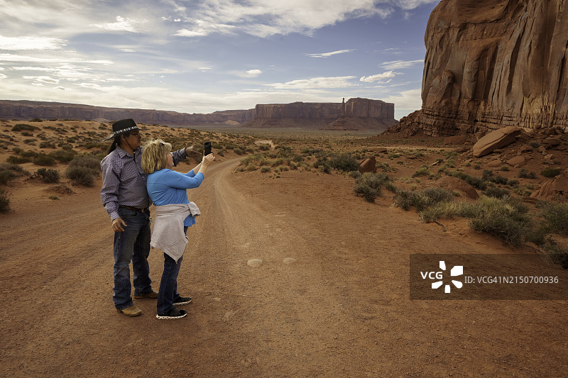 男性导游在纪念碑谷部落公园犹他州帮助女游客用智能手机拍摄著名地标的壮丽日落照片图片素材