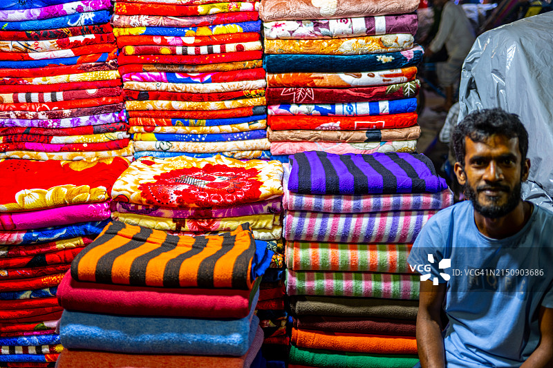 达卡市场景象：一位商人笑容满面地站在一堆色彩鲜艳的毯子中图片素材
