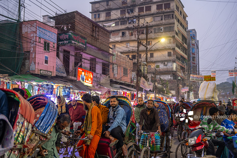 达卡夜景：黄昏时分，人力车夫在繁华的街道上排队，当地商店在纵横交错的电缆下发出温暖的光芒，捕捉了这座城市的活力。图片素材