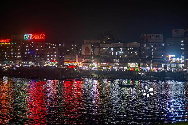 达卡萨达尔 घाट夜景：充满活力的灯光倒映在水面上，一艘孤零零的船漂过灯火通明的商店和霓虹灯，捕捉了这座城市热闹的河滨景象。图片素材