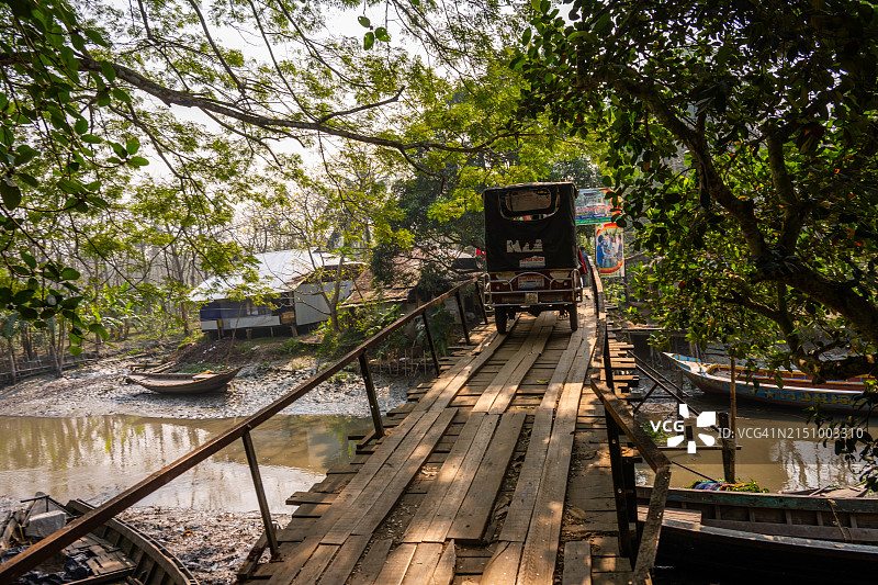 孟加拉国内萨拉巴德，人力车穿过安静的河流上的旧木桥，河岸边有船只和郁郁葱葱的阳光斑驳的树木。图片素材