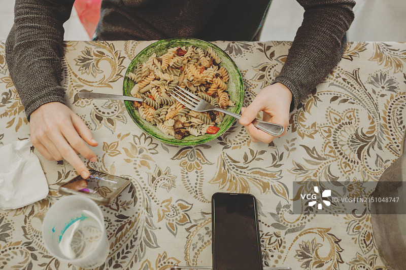 年轻男子用智能手机吃一碗蔬菜通心粉图片素材