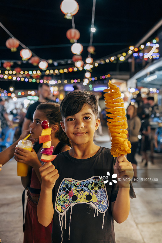 在食品市场上手持旋风薯条和水果串的微笑男孩肖像图片素材