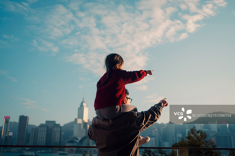 充满爱意的亚洲父亲扛着小女儿在肩上，指着远方，眺望着维多利亚港海滨长廊的城市天际线。享受父女共处的美好时光。家庭爱和团结的生活方式图片素材