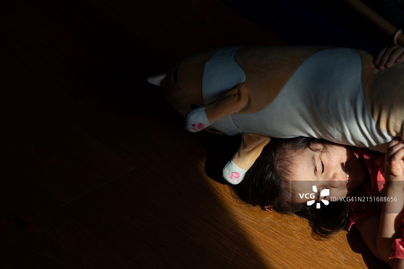 一个黑发女孩躺在地板上，旁边是一个大的毛绒玩具，沐浴在光线中并笑着图片素材