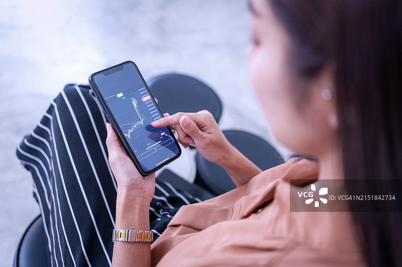 亚洲女人在家用智能手机监控加密货币和股票交易。图片素材