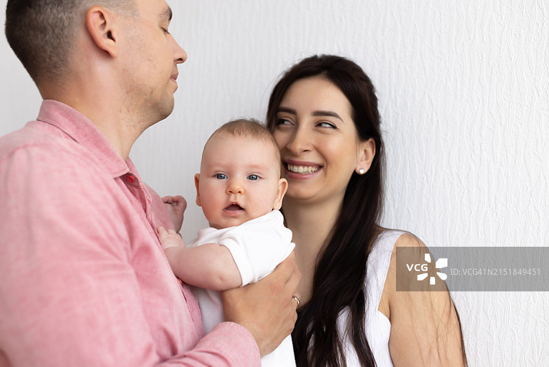 成年家庭，年轻父母站立并抱着3-4个月大的婴儿靠在白墙上图片素材