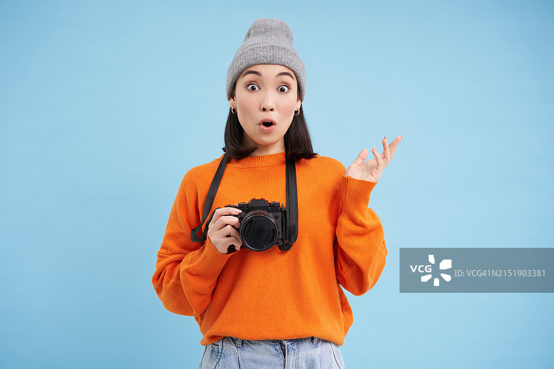 戴着帽子，拿着数码相机，看起来惊讶和印象深刻的惊讶女孩，在蓝色背景上摆姿势图片素材