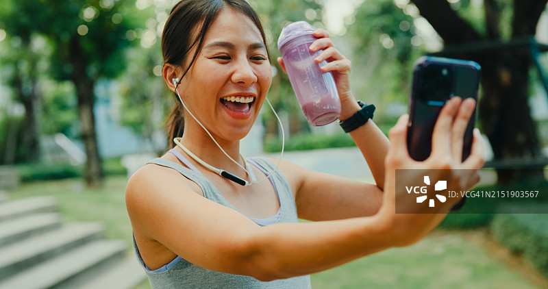 年轻的亚洲女运动员在城市公园锻炼后，对着智能手机挥手，与健身教练进行在线视频通话。健康的锻炼图片素材