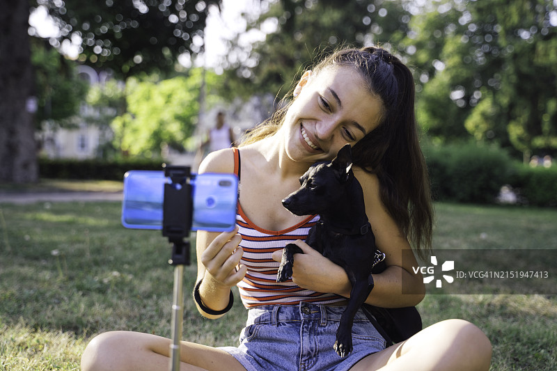笑容灿烂的少女在公园里用手机拍摄宠物狗Vlog，网红录制视频图片素材