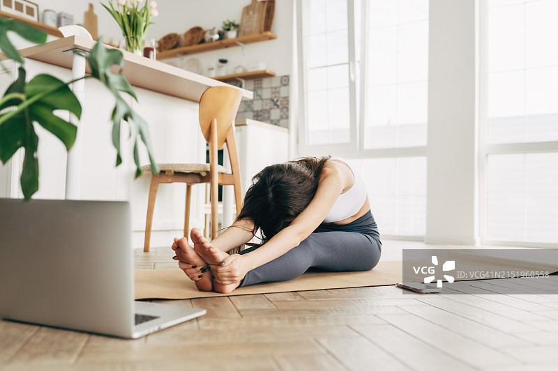 穿着运动装的健壮女人在家使用笔记本电脑在运动垫上做瑜伽图片素材