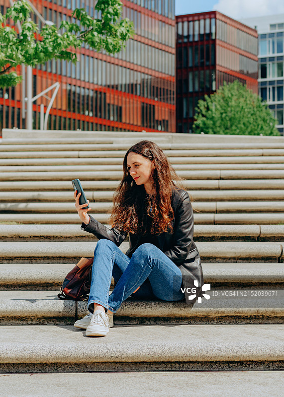 快乐的年轻女商人坐在台阶上用智能手机查看电子邮件图片素材