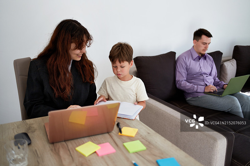 年轻父母在家使用笔记本电脑工作，儿子向母亲展示笔记本上的画作。工作与生活平衡概念。图片素材