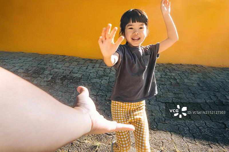 父亲向朝他跑来的可爱小女儿伸出手，父女在黄色墙壁前的路面上互动的视角。父女亲情时刻。家庭爱和团聚生活方式图片素材