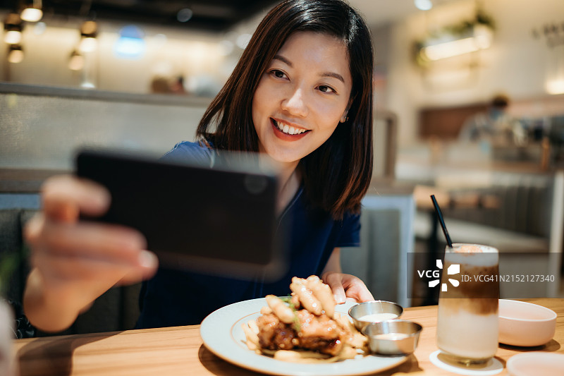 微笑的亚洲年轻女子在咖啡馆享用甜点时用智能手机自拍，并在社交媒体上分享时光。相机先吃。青年文化。有趣的生活方式和科技图片素材