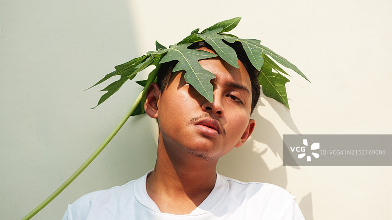 英俊的亚洲年轻男人特写，面带倦容，头顶木瓜叶图片素材