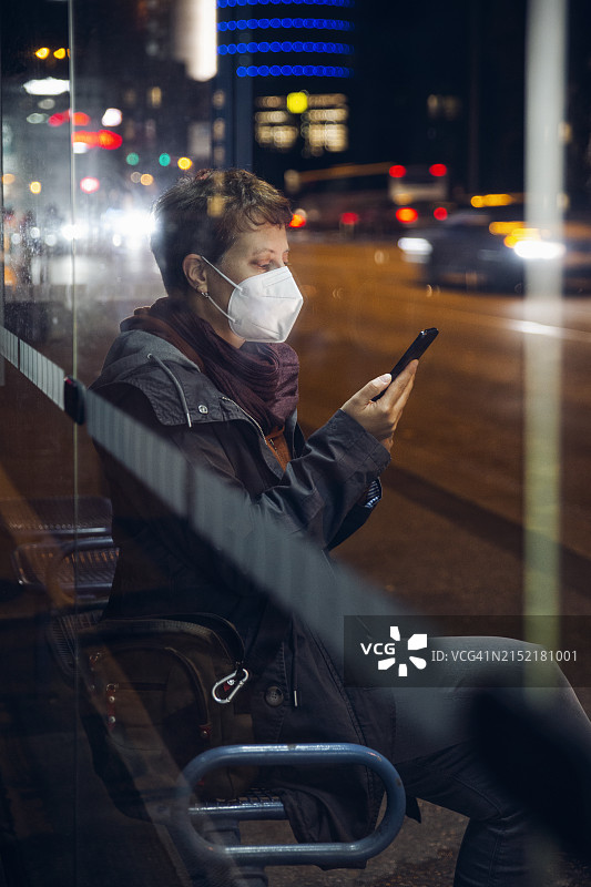 戴防护口罩的中年妇女在城市公交车站使用手机图片素材