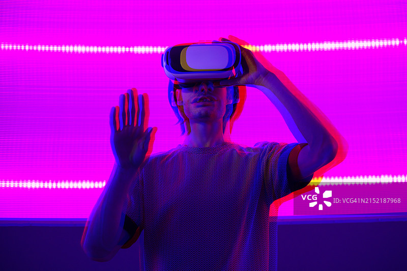 惊讶的年轻男子戴着虚拟现实眼镜，在蓝色霓虹灯墙背景上手势图片素材