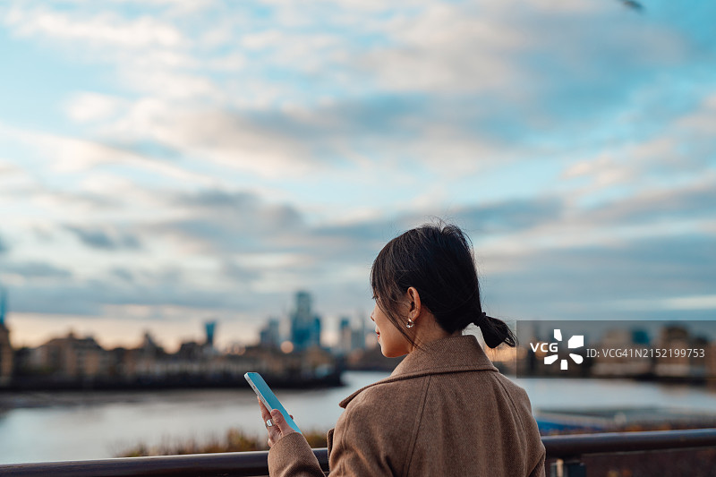 在城市天际线下使用智能手机的年轻亚洲女子背影图片素材