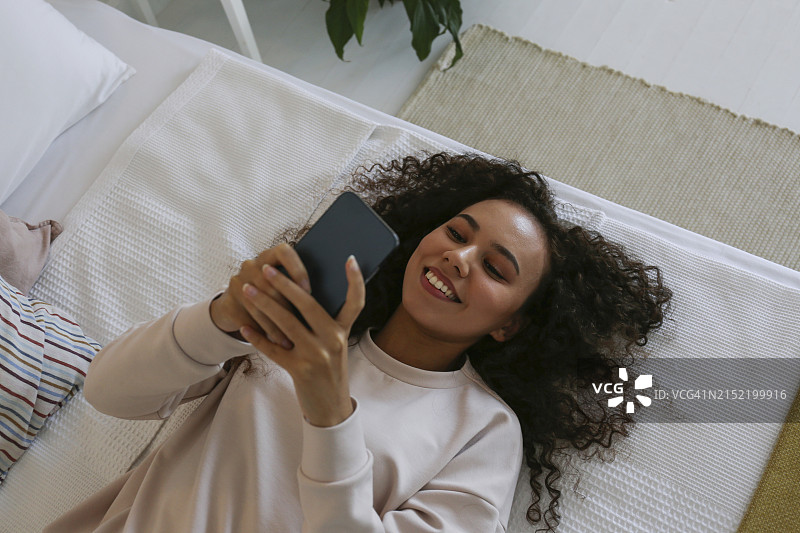 快乐的年轻女人躺在床上，对着手机微笑图片素材