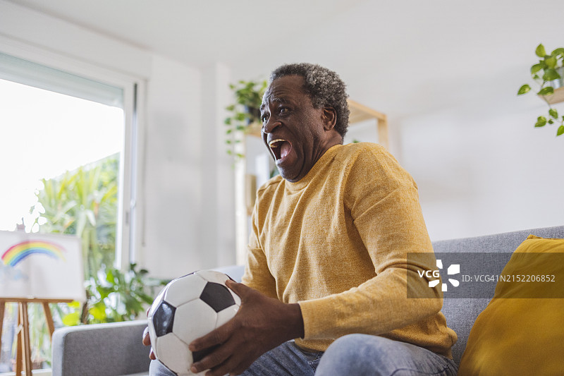 快乐的老年男人抱着足球在家观看比赛图片素材