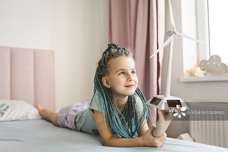 笑容灿烂的女孩染着辫子发型，在家躺在床上拿着风力涡轮机模型图片素材