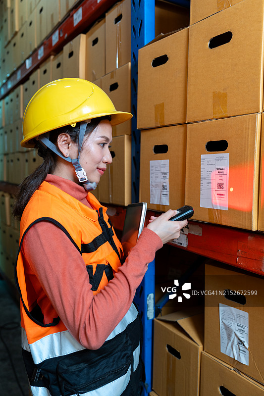 美丽的亚洲女人戴着黄色头盔和反光橙色安全背心，使用无线条形码阅读器扫描贴纸条形码，以验证仓库中产品包装盒的真伪图片素材