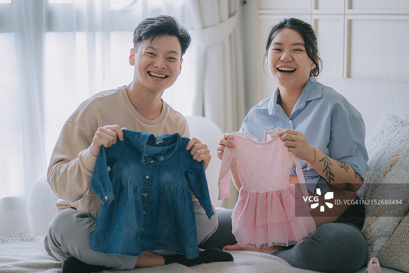 快乐的年轻亚裔中国怀孕夫妇拿着女婴服装，看着镜头微笑坐在床上图片素材