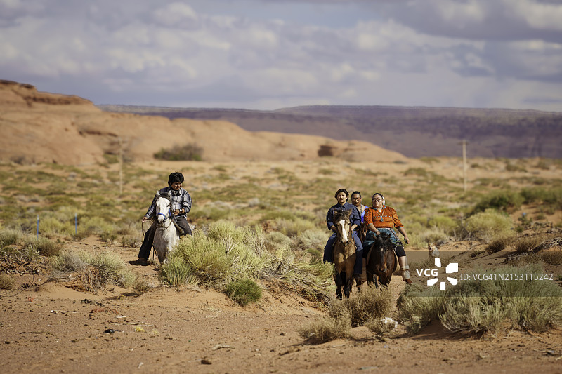 年轻的纳瓦霍族家庭在纪念碑谷亚利桑那州牧场上骑马，背景是壮丽的红色岩石山脉和引人注目的云景图片素材