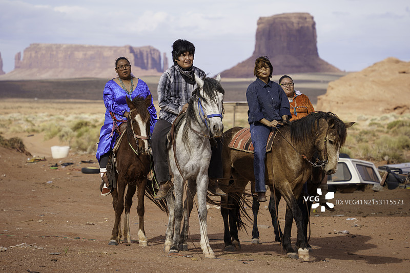 年轻的纳瓦霍族家庭在纪念碑谷亚利桑那州牧场上骑马，背景是壮丽的红色岩石山脉和引人注目的云景图片素材