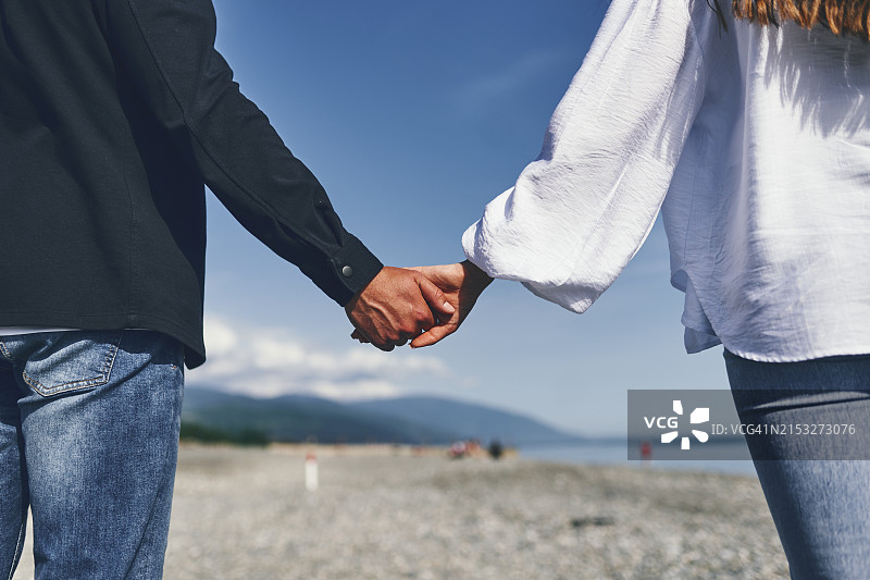 情侣在海滩上手牵着手，穿着休闲装，背景是鹅卵石海滩、蓝天和群山图片素材