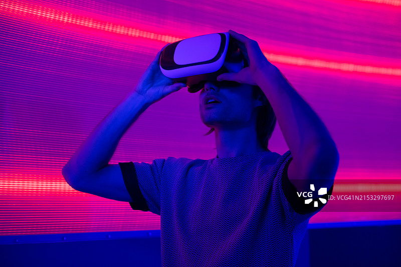 惊讶的创意男士戴着VR眼镜，在蓝色霓虹灯墙背景上手势比划图片素材