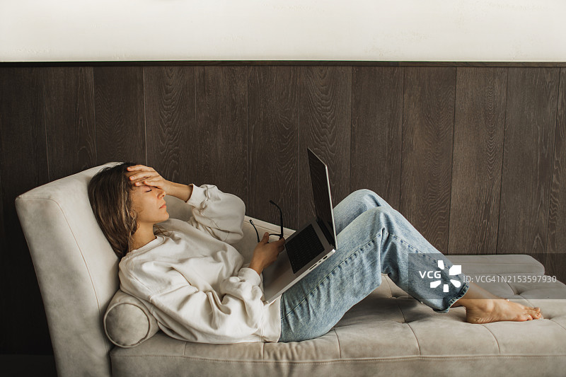 工作后，疲惫的女性因头痛躺在沙发上用笔记本电脑图片素材