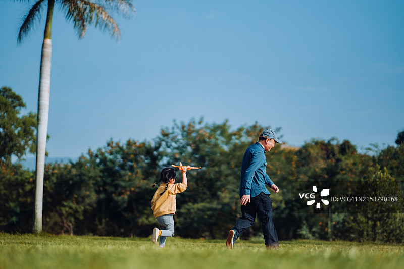 亚洲爷爷和可爱的小孙女在户外共度时光，在美丽的蓝天下，在公园里玩飞机玩具，快乐地微笑。多代家庭。爱与团结的概念图片素材