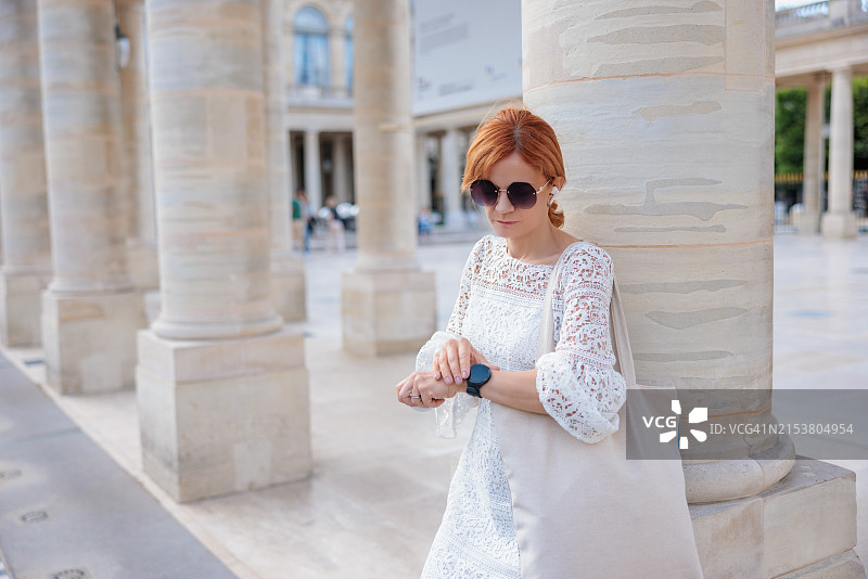 戴着太阳镜站在建筑圆柱旁的红发女子，正在查看她的智能手表图片素材