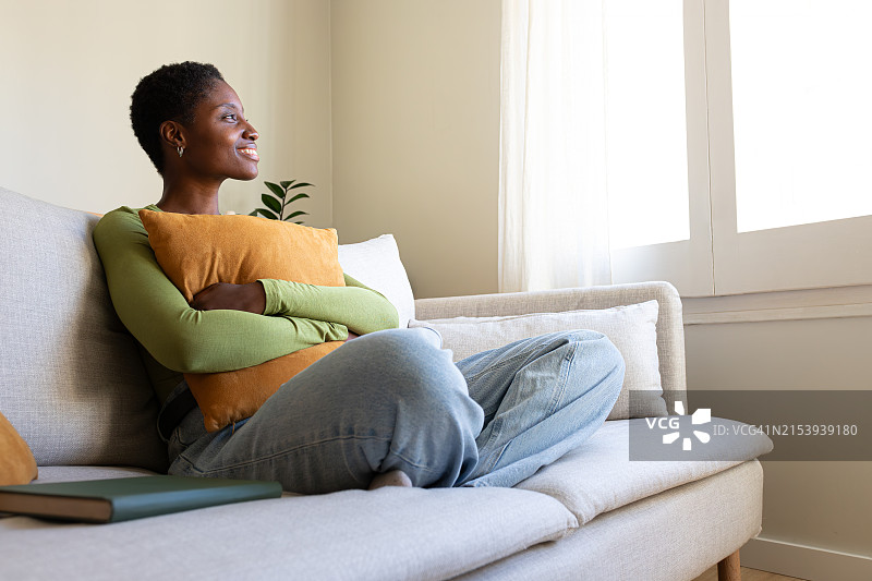 年轻沉思的非裔美国女人在家放松，面带微笑地望着窗外侧视图。留白图片素材