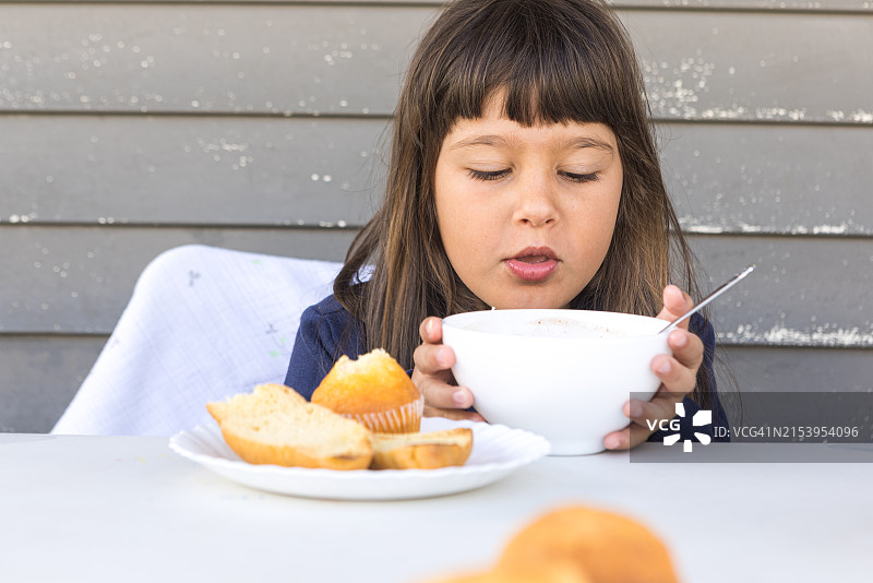 黑发女孩在户外餐桌旁吃早餐， Concept of innocence, breakfast, family, naturalness and routine。图片素材