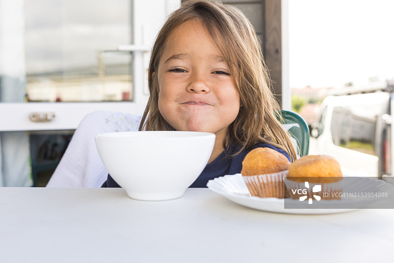 金发女孩在户外餐桌旁吃早餐，对着碗和盘子里的两个松饼微笑，象征着天真、早餐、家庭、自然和规律。图片素材
