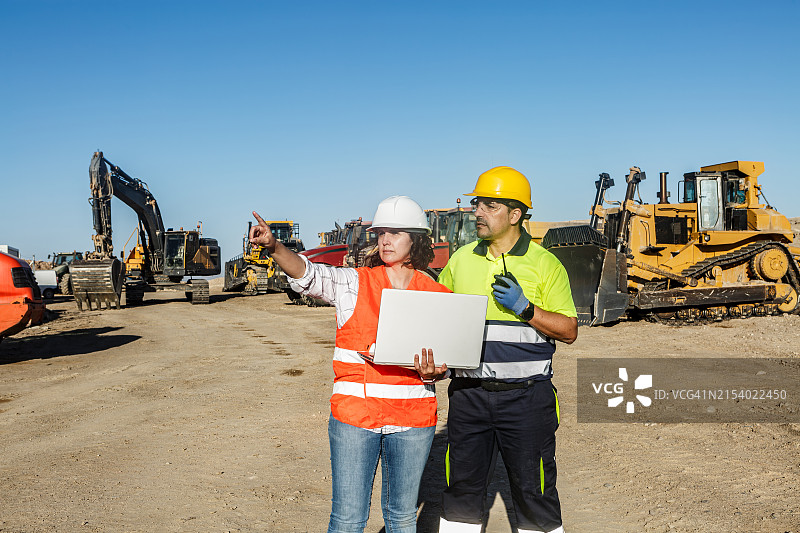 阳光明媚的日子里，建筑工人审查带有重型机械的现场平面图图片素材
