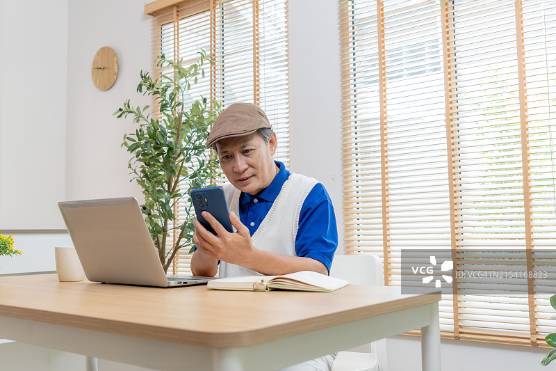 在家休闲时用手机与妻子通话发短信的年长亚洲男人。沉迷社交媒体，享受科技带来的快乐的年长男人。退休。图片素材