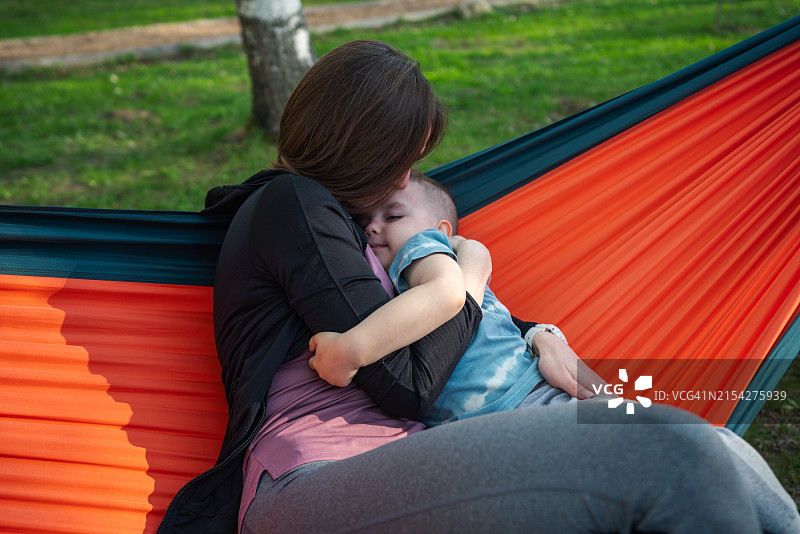 户外吊床上，母亲拥抱亲吻小儿子图片素材