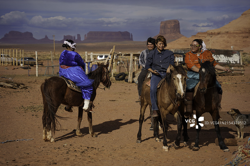 年轻的纳瓦霍族家庭在纪念碑谷亚利桑那州牧场上骑马，背景是壮丽的红色岩石山脉和引人注目的云景图片素材