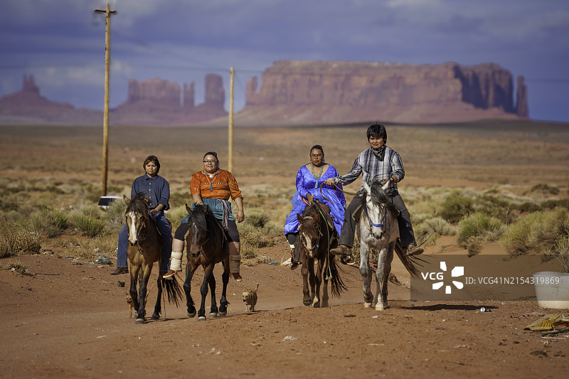 年轻的纳瓦霍族家庭在纪念碑谷亚利桑那州牧场上骑马，背景是壮丽的红色岩石山脉和引人注目的云景图片素材