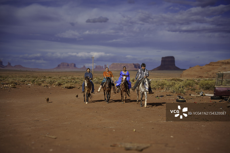年轻的纳瓦霍族家庭在纪念碑谷亚利桑那州牧场上骑马，背景是壮丽的红色岩石山脉和引人注目的云景图片素材