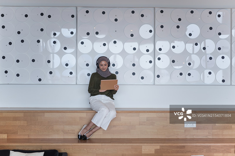一位戴着头巾和耳机，坐在现代室内空间的木凳上阅读平板电脑的女人图片素材