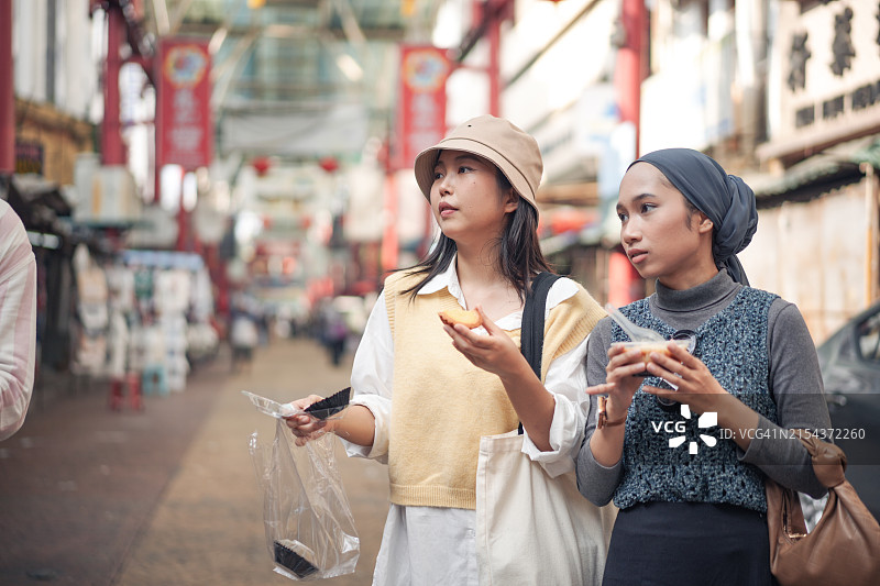 两位年轻女人在热闹的户外市场品尝街头小吃图片素材