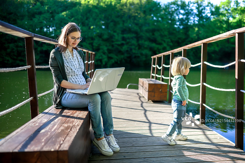 在湖边用笔记本电脑工作：戴眼镜的妈妈和幼儿在公园里享受亲子时光，远程工作和育儿图片素材